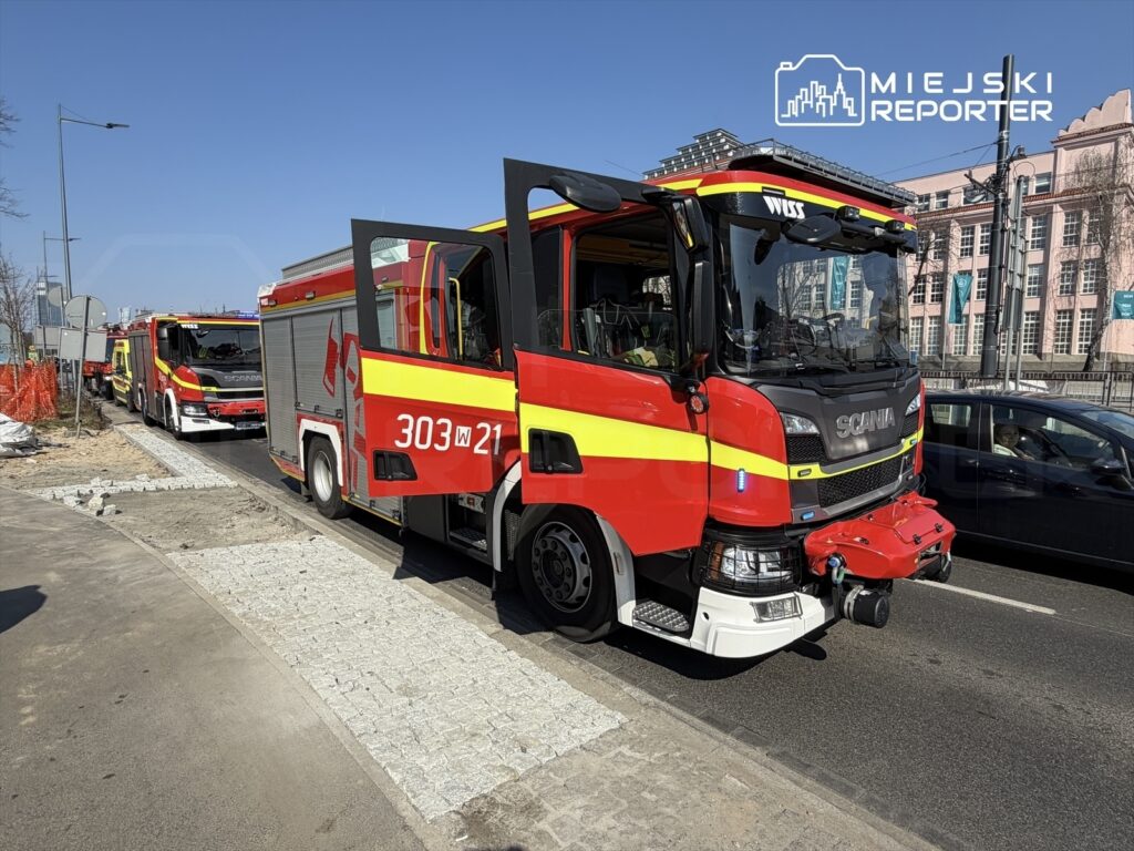 Czerwony wóz strażacki marki Scania zatrzymany na ulicy, obok niego drugi wóz i widok na nowoczesne budynki w tle.