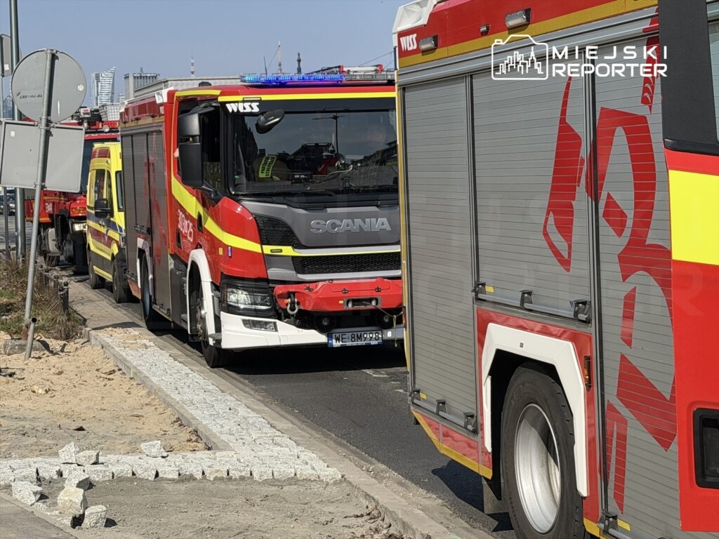 Czerwone pojazdy straży pożarnej ustawione w kolejce na ulicy, w tle nowoczesne budynki.
