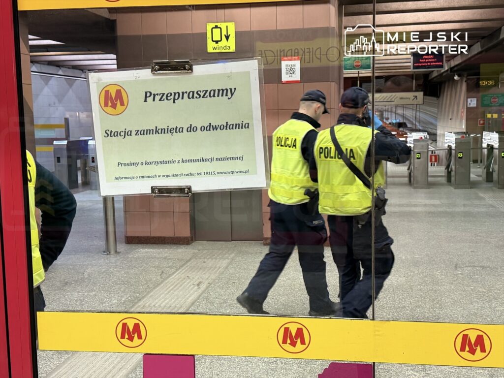 Dwaj policjanci w kamizelkach odblaskowych rozmawiają przy zamkniętej stacji metra, obok widoczny komunikat o zamknięciu.