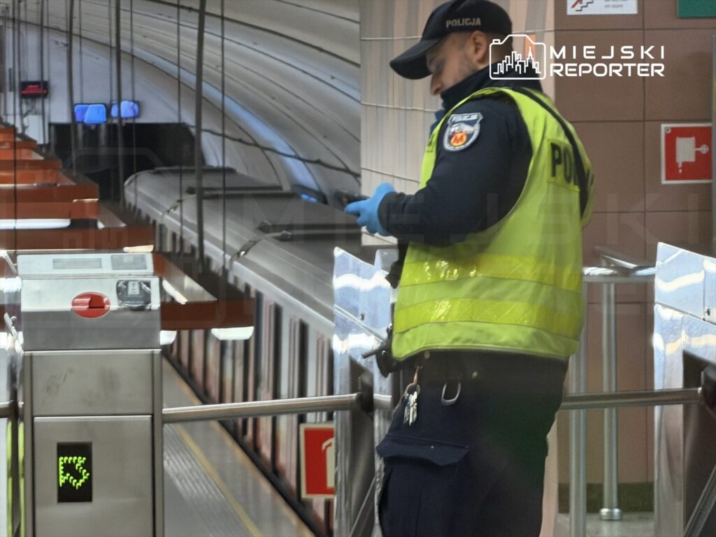 Policjant w odblaskowej kamizelce sprawdza telefon na stacji metra, obok zamkniętego pociągu.