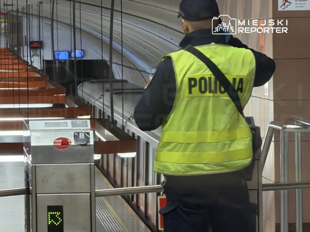 Policjant w żółtej kamizelce kontroluje przejście na stacji metra, w tle widoczne wagony pociągu.