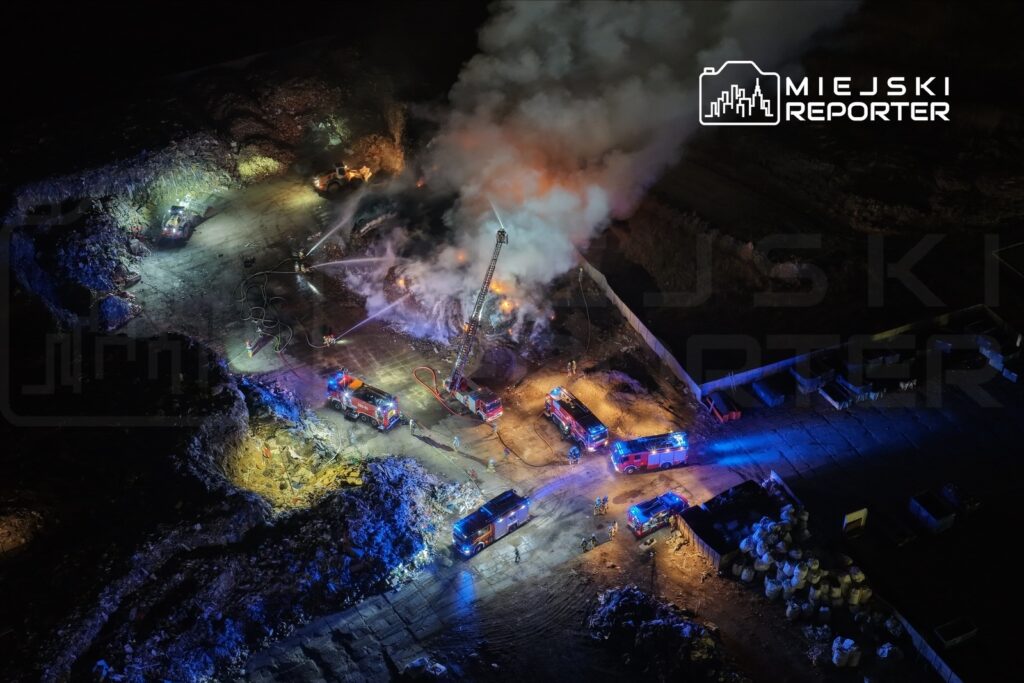 Grupa strażaków gasi pożar na terenie przemysłowym, otoczonym przez pojazdy gaśnicze i unoszący się dym.