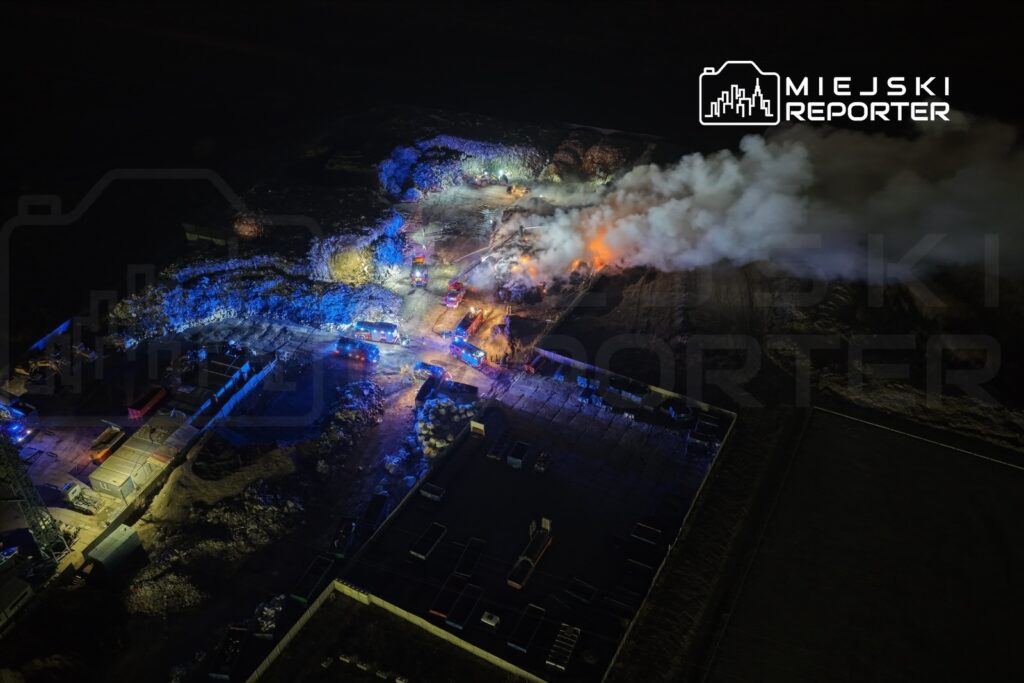 W nocy strażacy gaszą pożar na terenie składowiska, otoczonym przez niebieskie światła pojazdów ratunkowych.