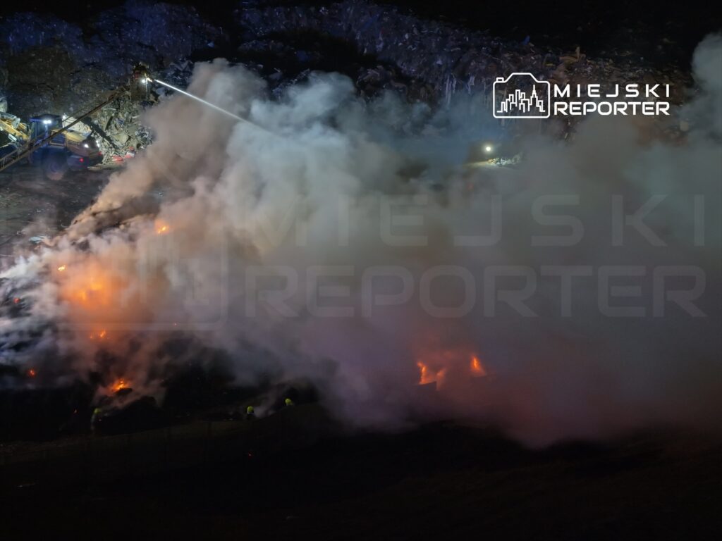 Strażacy gaszą pożar na wysypisku, otoczeni gęstym dymem i płomieniami, w nocy.