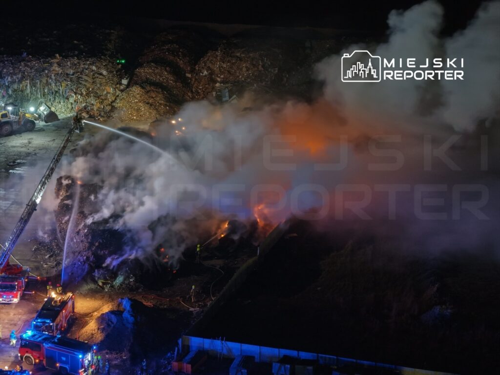 Strażacy gaszą pożar na wysypisku, używając węży strażackich, przy intensywnym zadymieniu i płomieniach.