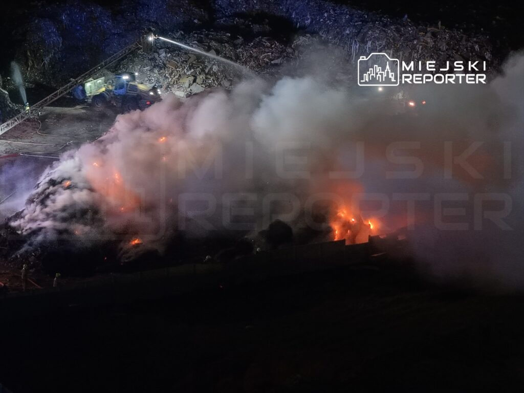 Ogień wydobywający się z hałdy odpadów, gęsty dym unoszący się w powietrzu, w tle widoczny sprzęt gaśniczy.