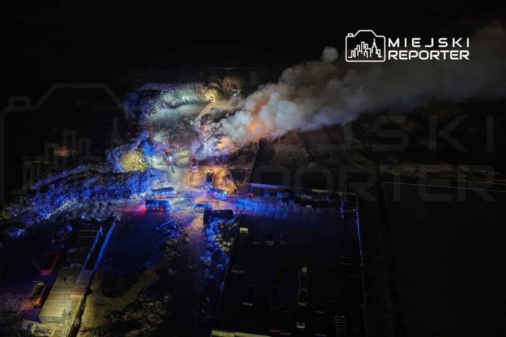 W nocy strażacy gaszą pożar w terenie przemysłowym, otoczonym przez pojazdy ratunkowe i dym.