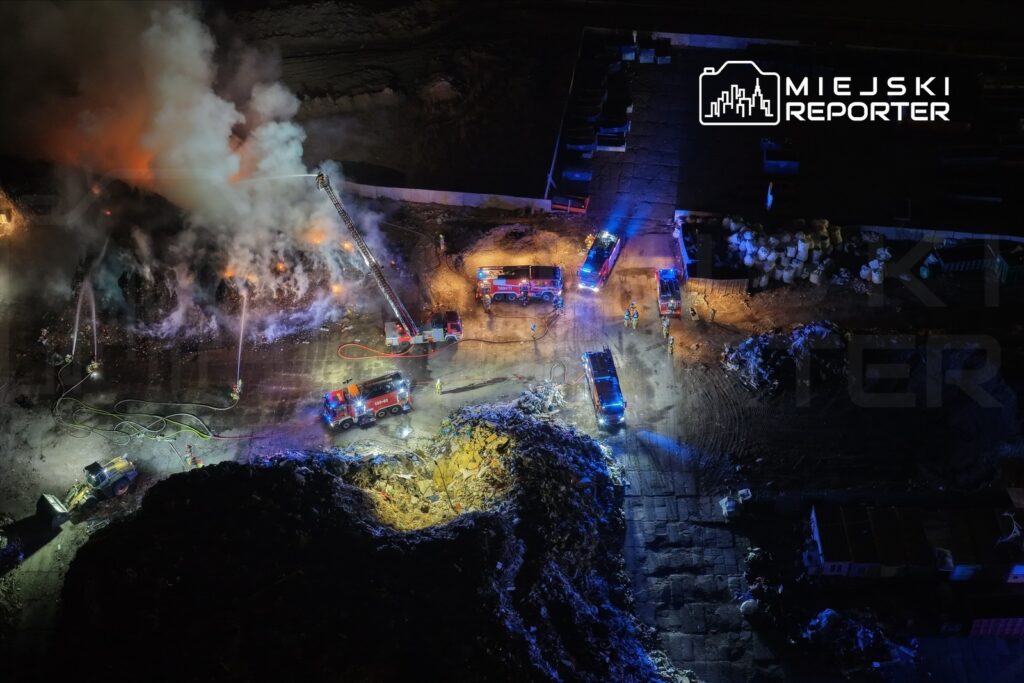 Strażacy gaszą pożar na wysypisku śmieci, używając węży strażackich i sprzętu gaśniczego w nocy.