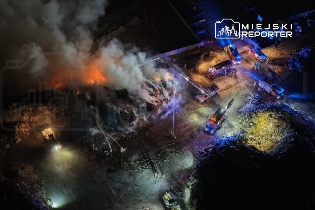 Strażacy gaszą pożar na wysypisku śmieci, otoczeni dymem i płomieniami, z widocznymi wozami strażackimi.