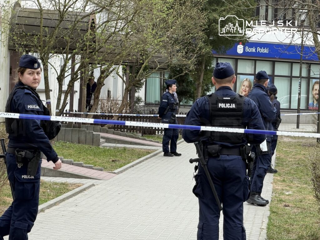 Grupa policjantów w mundurach stoi na chodniku, zabezpieczając teren taśmą policyjną przed budynkiem banku.
