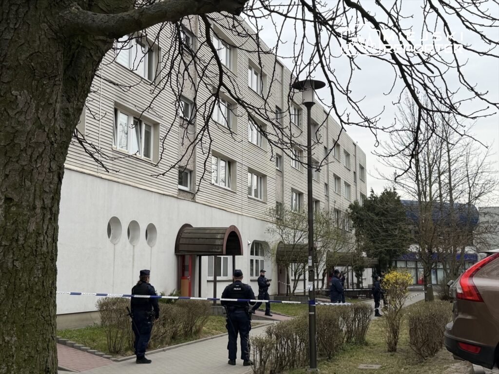 Grupa policjantów zabezpiecza teren przed budynkiem mieszkalnym, obok stoją zaparkowane samochody.