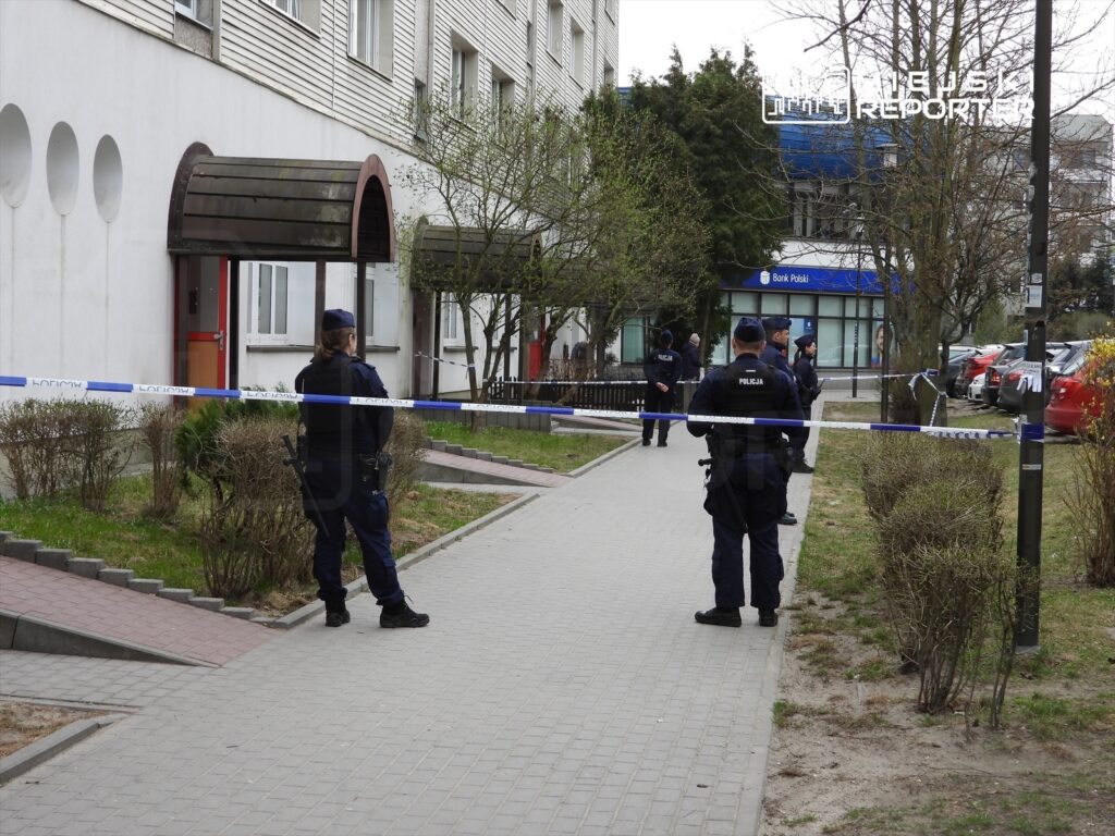 Policjanci zabezpieczają teren przed budynkiem mieszkalnym, na którym widać niebieską taśmę policyjną.