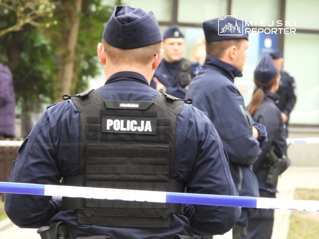Policjant w kamizelce z napisem "POLICJA" stoi za taśmą zabezpieczającą, obserwując innych funkcjonariuszy.