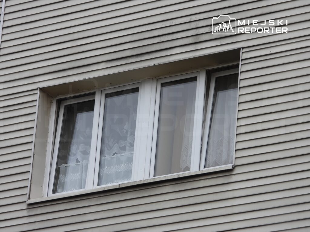 Okna z białymi ramami, z firankami, umieszczone w elewacji budynku z poziomymi panelami.