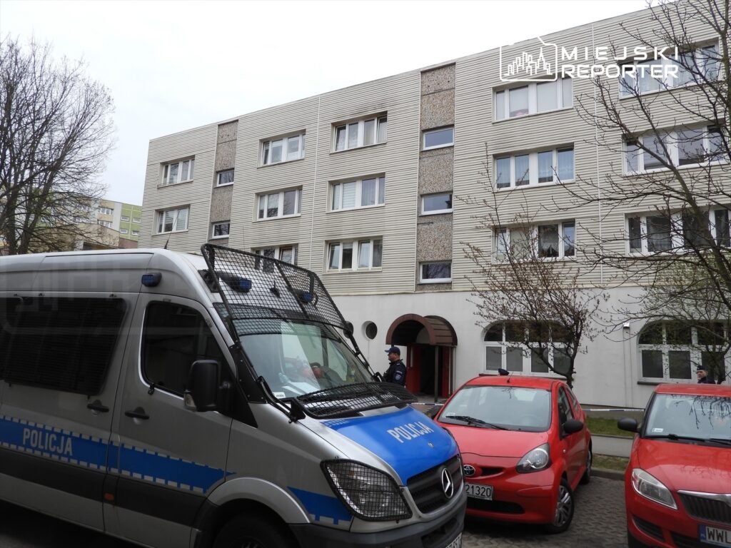 Policyjny radiowóz zaparkowany przed budynkiem mieszkalnym, obok czerwone samochody osobowe, w tle drzewa.