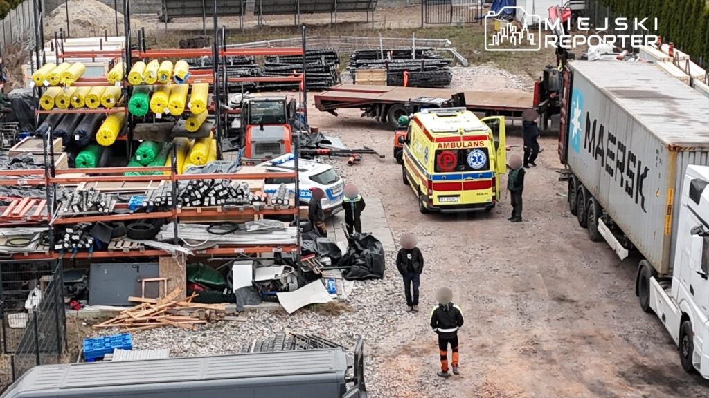 Na placu budowy kilka osób rozmawia obok karetki pogotowia, a w tle widoczne są materiały budowlane na regałach.