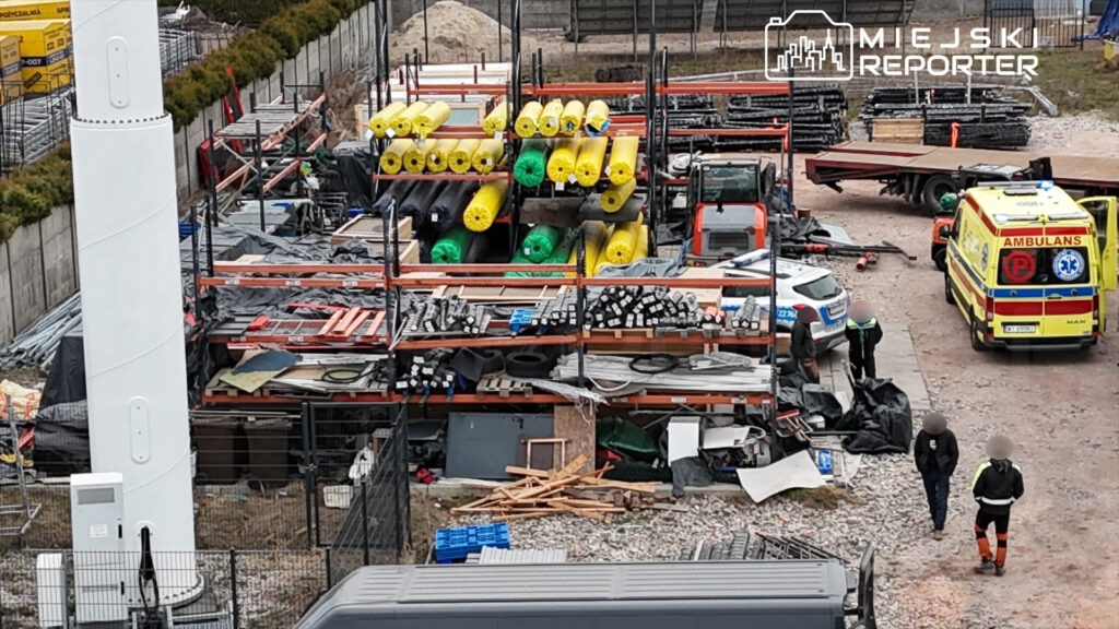 Na placu budowy widać zorganizowane materiały budowlane, w tym rolki z tworzywa sztucznego i metalowe rury. Obok parkują pojazdy, w tym karetka oraz radiowóz, a pracownicy rozmawiają w grupach.