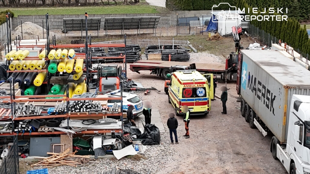 Na placu budowy zespół pracowników otacza karetkę, obok stoją ciężarówki i sprzęt budowlany.
