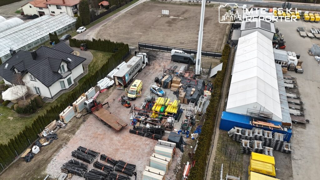 Teren budowy z różnorodnym sprzętem i materiałami, w tym ciężarówkami oraz ambulansami, otoczony zielonymi żywopłotami.