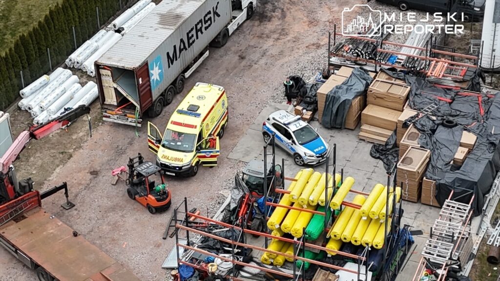 Karetka pogotowia oraz radiowóz policyjny stoją na placu budowy otoczonym materiałami budowlanymi i kontenerami.