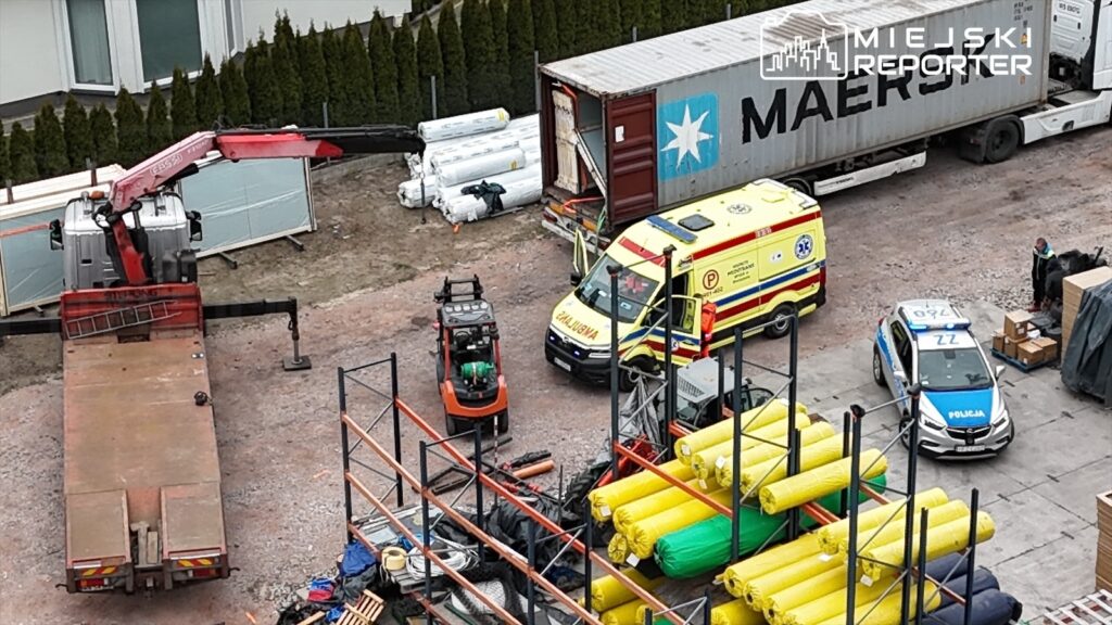 Na placu budowy stoi wózek widłowy obok karetki pogotowia i radiowozu policji, w tle kontener transportowy.