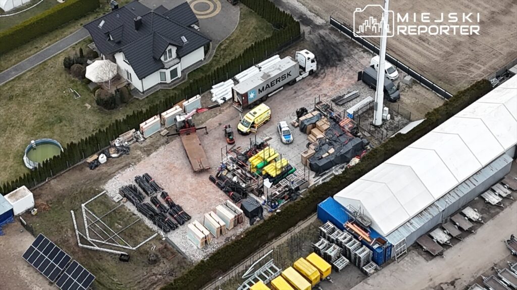 Na placu budowy znajdują się maszyny i materiały budowlane, a obok widoczny jest samochód dostawczy oraz karetka.