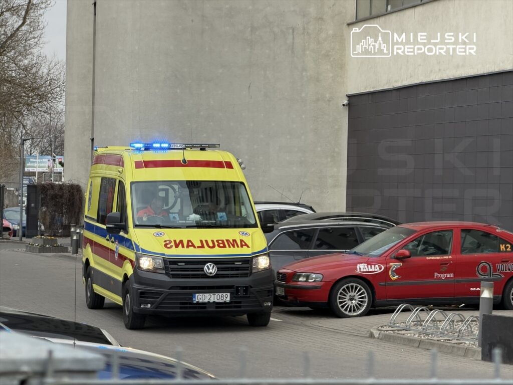 Karetka pogotowia z włączonymi sygnałami świetlnymi wjeżdża na parking obok zaparkowanych samochodów.
