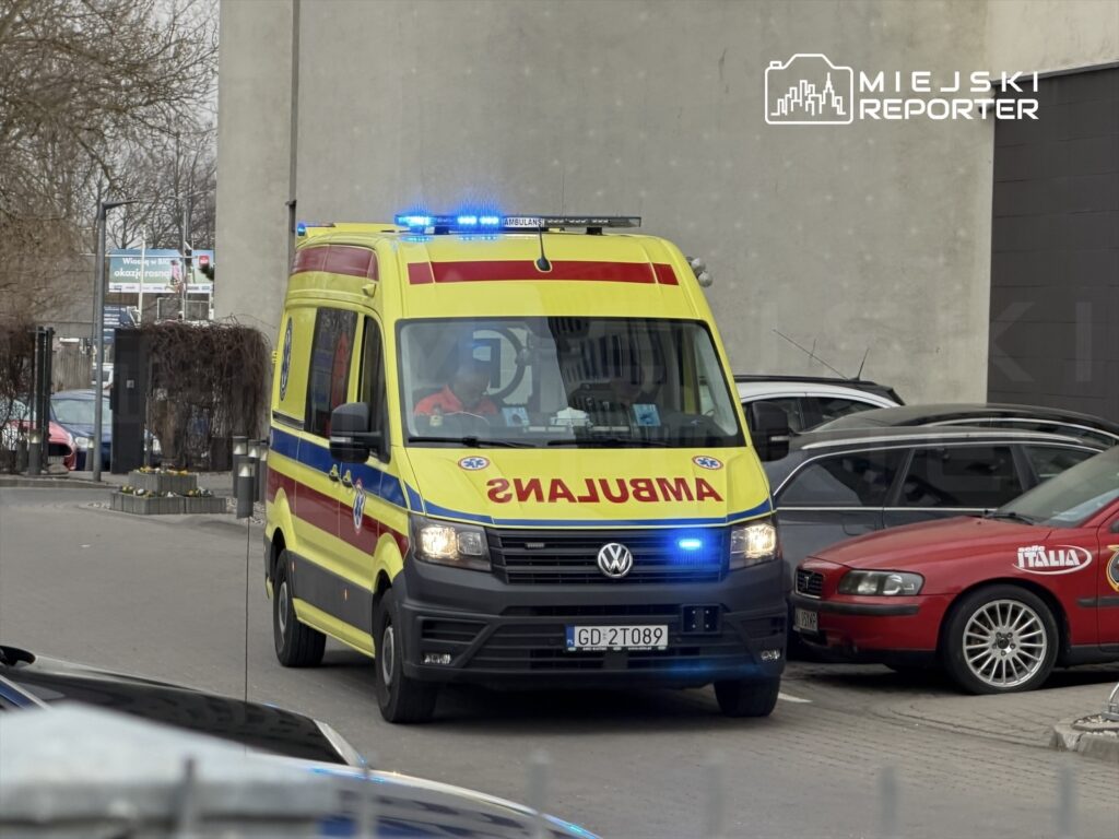 Ambulans z włączonymi sygnałami świetlnymi wjeżdża na parking w miejskim otoczeniu.