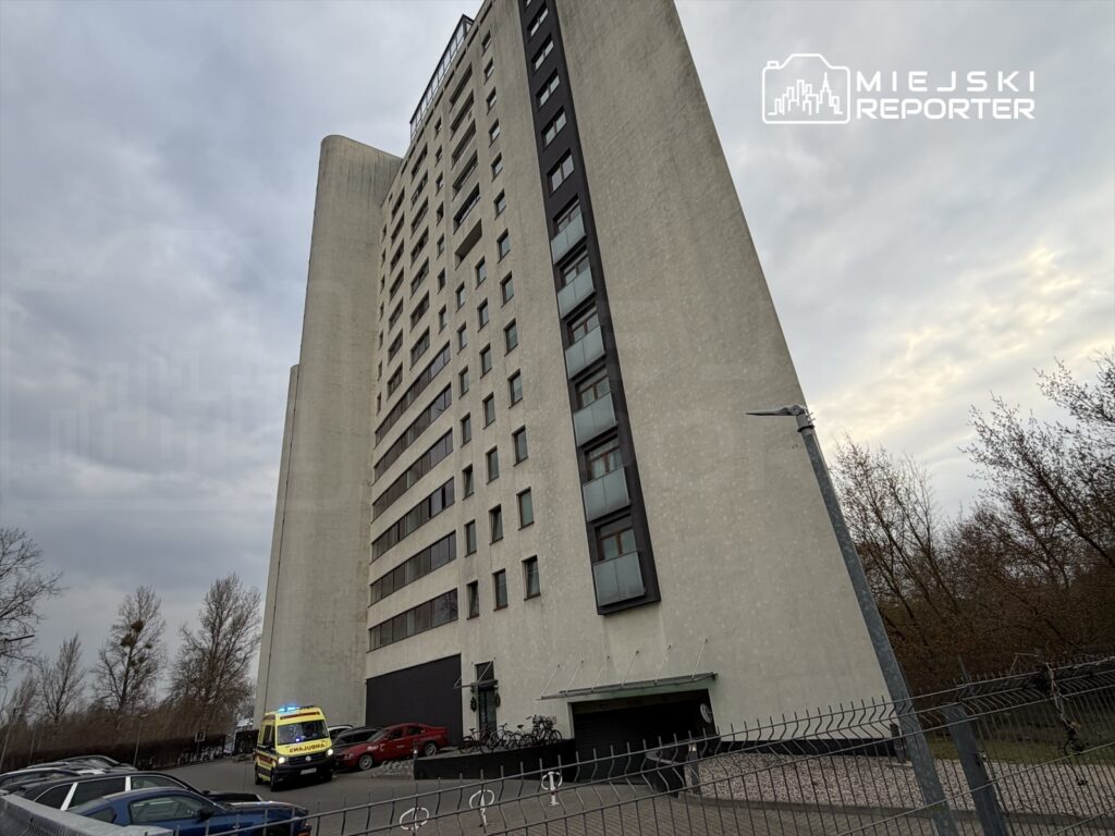 Wysoki budynek mieszkalny z szarym tynkiem i dużymi oknami, obok zaparkowana karetka pogotowia.