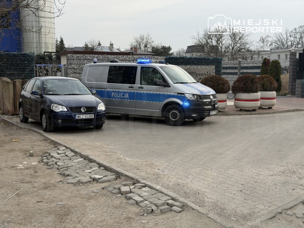 Policjant w niebieskim radiowozie z włączonymi sygnałami świetlnymi stoi obok zaparkowanego samochodu osobowego.