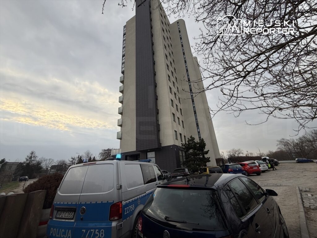 Policyjny radiowóz z włączonymi sygnałami świetlnymi zaparkowany obok nowoczesnego budynku mieszkalnego.