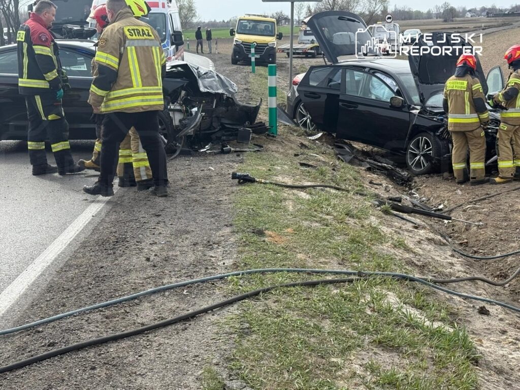 Grupa strażaków ocenia skutki wypadku drogowego, przy uszkodzonym pojeździe na poboczu drogi.