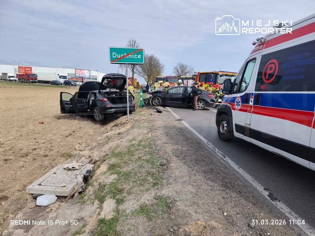 Na drodze stoi rozbity samochód, obok niego zespół ratunkowy oraz karetka, w tle znak drogowy z nazwą miejscowości.