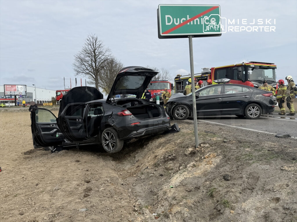 W miejscu wypadku drogowego, uszkodzony czarny samochód stoi na poboczu, a obok pracują strażacy.