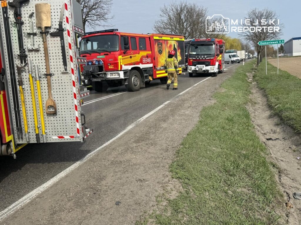 Strażacy w mundurach pracują przy dwóch wozach strażackich na poboczu drogi, obok znaku kierunkowym.