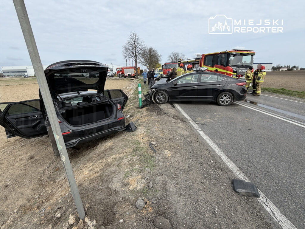 Czarny samochód osobowy zderzony z innym pojazdem na drodze, obok strażacy i służby ratunkowe.