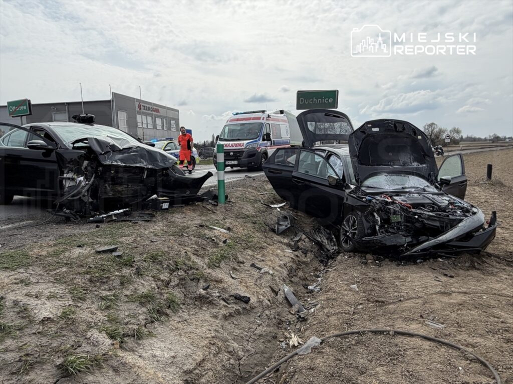 Dwa uszkodzone samochody osobowe na poboczu drogi, obok stoi zespół ratunkowy oraz znak drogowy "Duchnice".