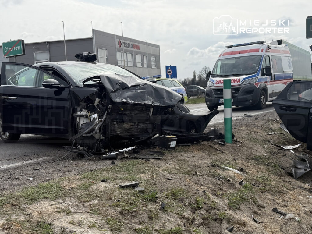 Uszkodzony samochód osobowy stoi na poboczu drogi, obok przejeżdża karetka. W tle widoczne są budynki i znaki drogowe.