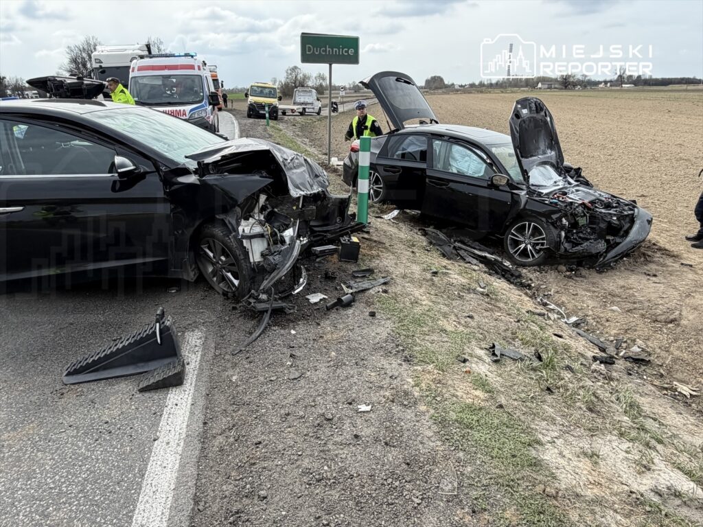 Dwa rozbite samochody osobowe na poboczu drogi, obok znaku miejscowości. Ratownicy medyczni udzielają pomocy poszkodowanym.