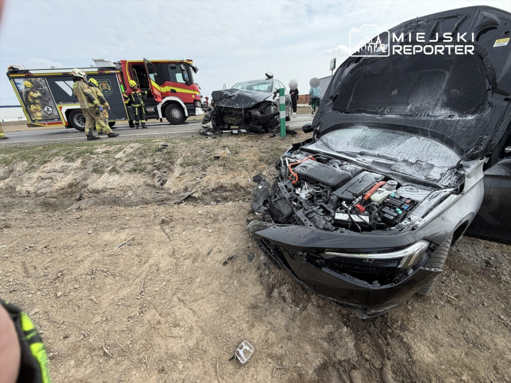 W miejscu wypadku drogowego strażacy pracują nad usunięciem rozbitego pojazdu, obok stoi wóz strażacki.