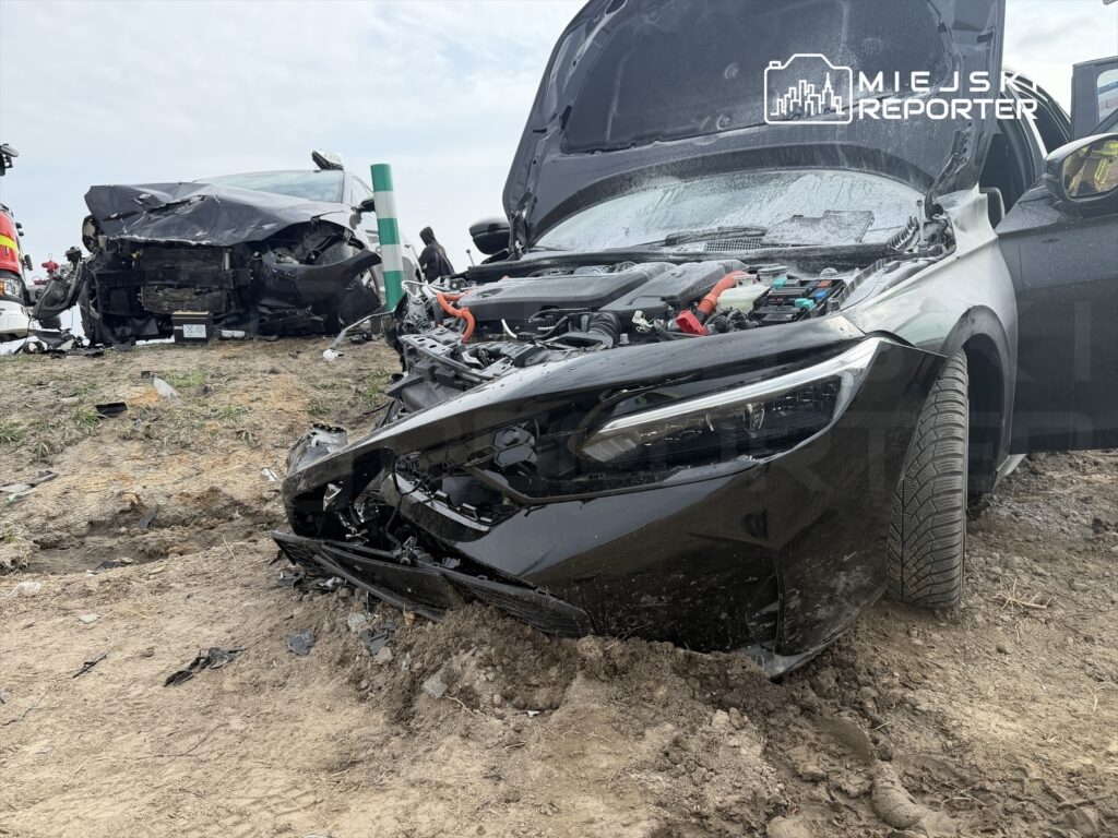 Czarny samochód osobowy z uszkodzoną przednią częścią stoi na poboczu drogi, obok widać drugi pojazd po wypadku.