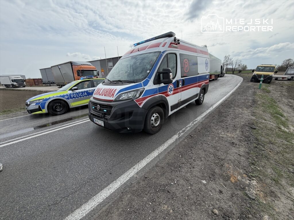 Karetka pogotowia i radiowóz policji zatrzymane na drodze obok parkujących ciężarówek w wiejskim otoczeniu.