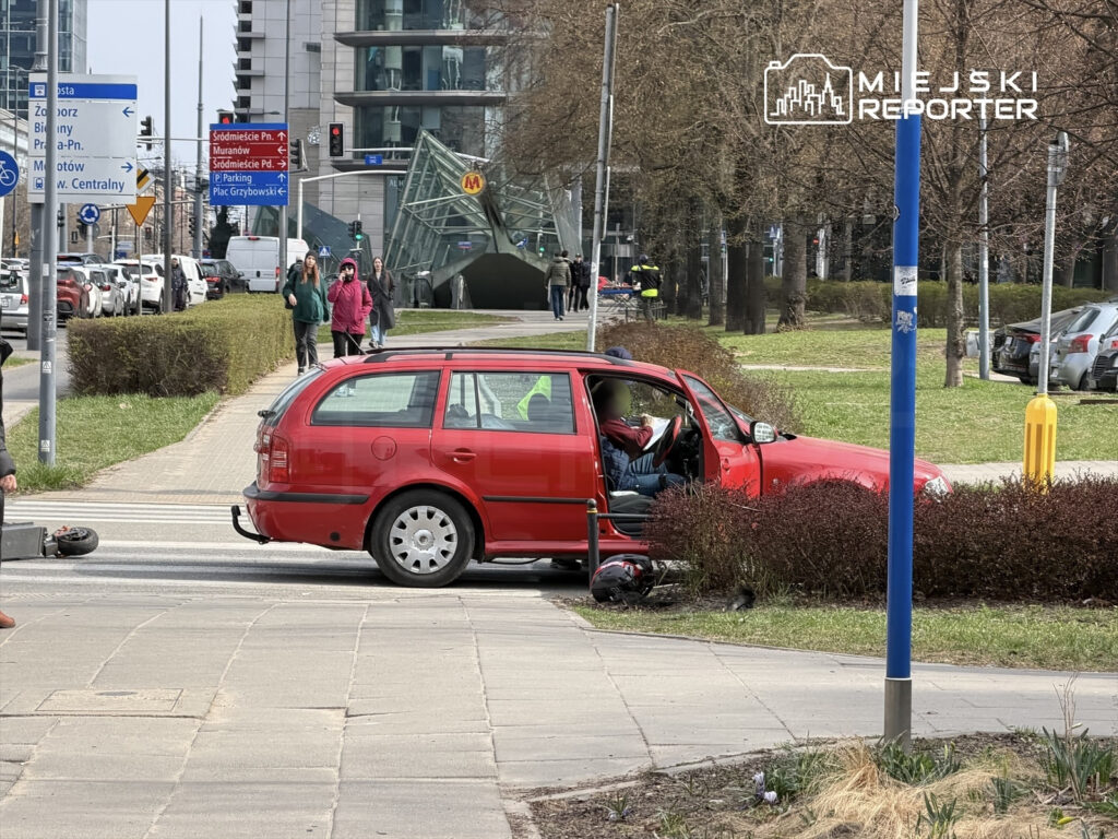 Czerwony samochód osobowy stoi na chodniku, a obok przechodzą piesi. W tle widać nowoczesne budynki i znaki drogowe.