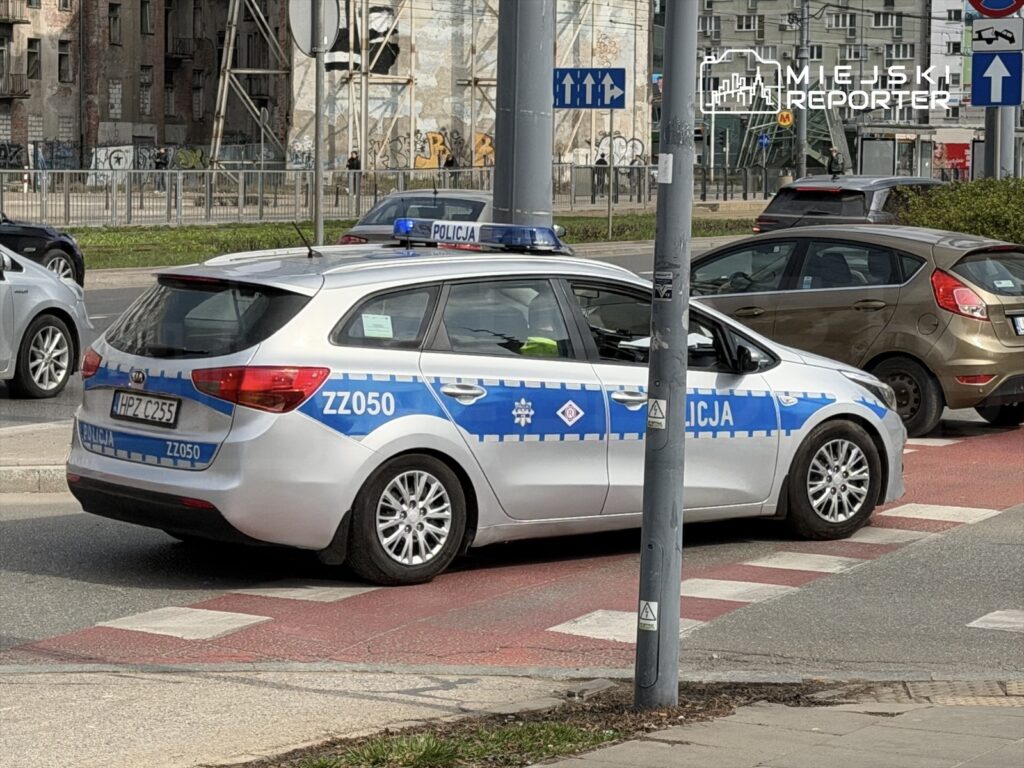 Policyjny samochód zaparkowany na skrzyżowaniu z oznaczeniami drogowymi w tle.