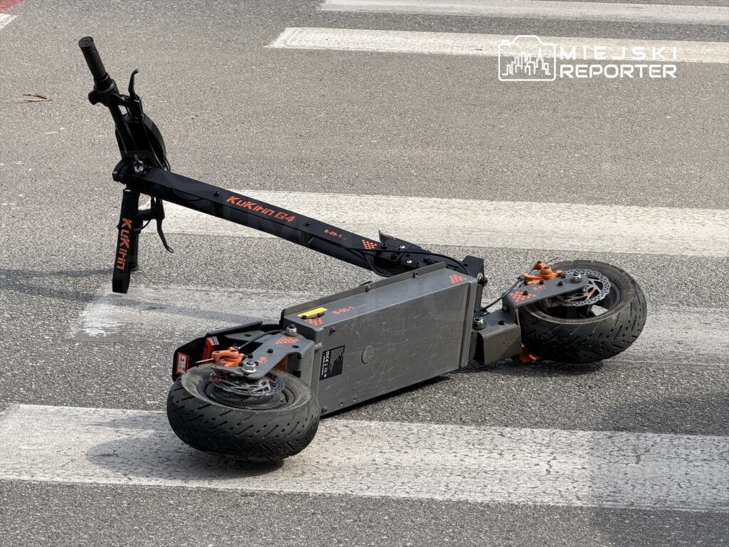 Złamana hulajnoga elektryczna leży na przejściu dla pieszych na asfaltowej ulicy.