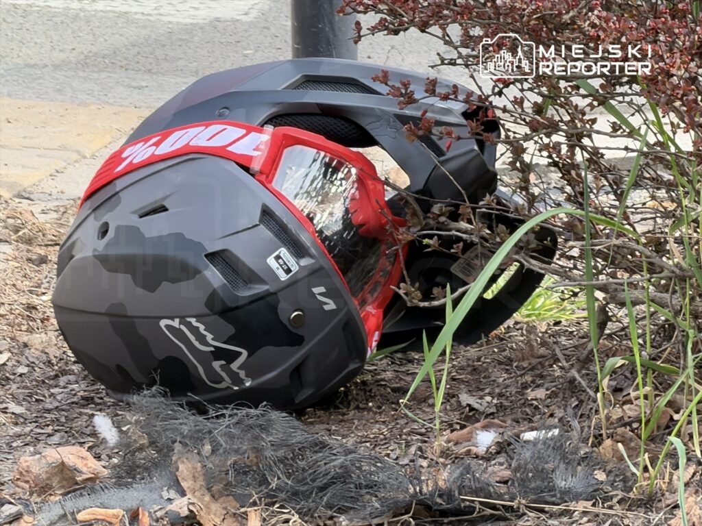 Czarny kask motocyklowy z czerwoną wstążką leży na ziemi obok krzewu, otoczony suchymi liśćmi i trawą.