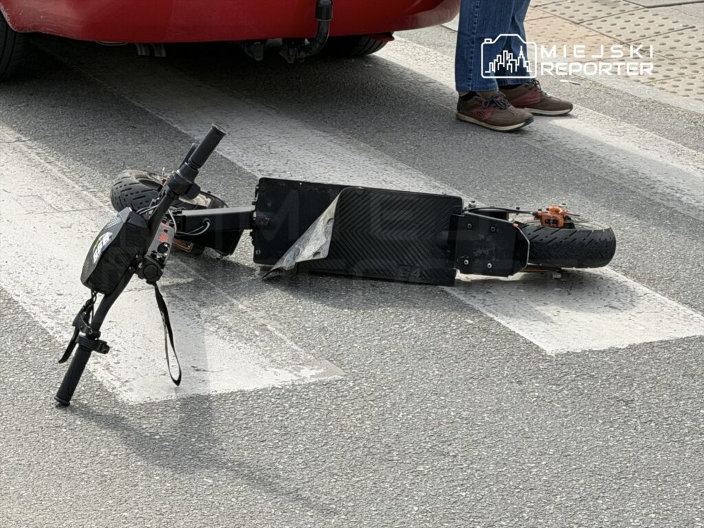 Uszkodzony skuter elektryczny leży na przejściu dla pieszych, obok czerwonego samochodu. W tle widoczna noga osoby w dżinsach.