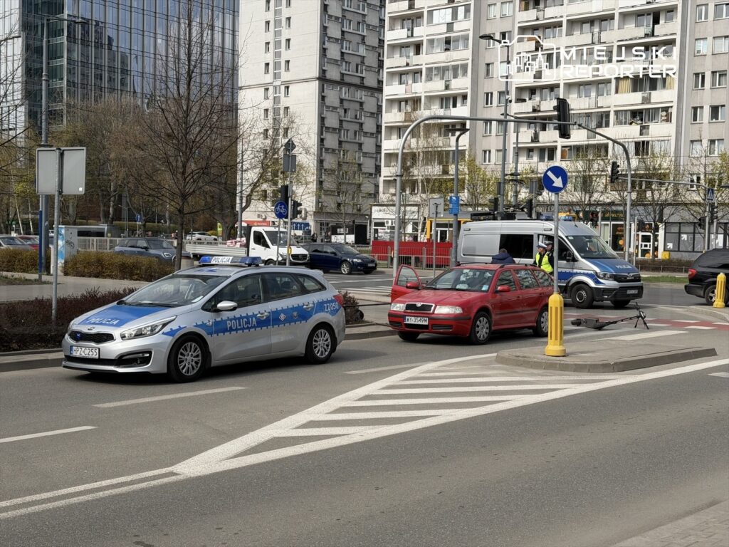Policyjny radiowóz zatrzymany na skrzyżowaniu, obok czerwona osobówka z otwartymi drzwiami, w tle nowoczesne budynki.