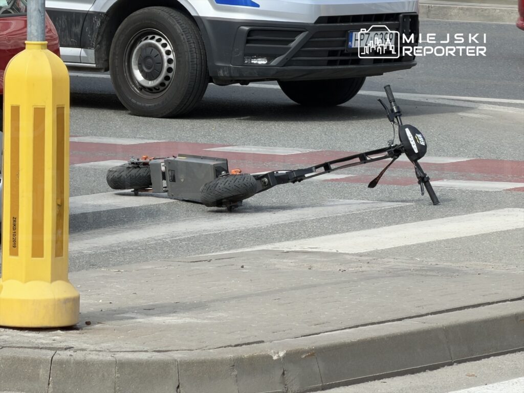 Na pasie drogowym leży przewrócona hulajnoga elektryczna, obok niej stoją samochody.