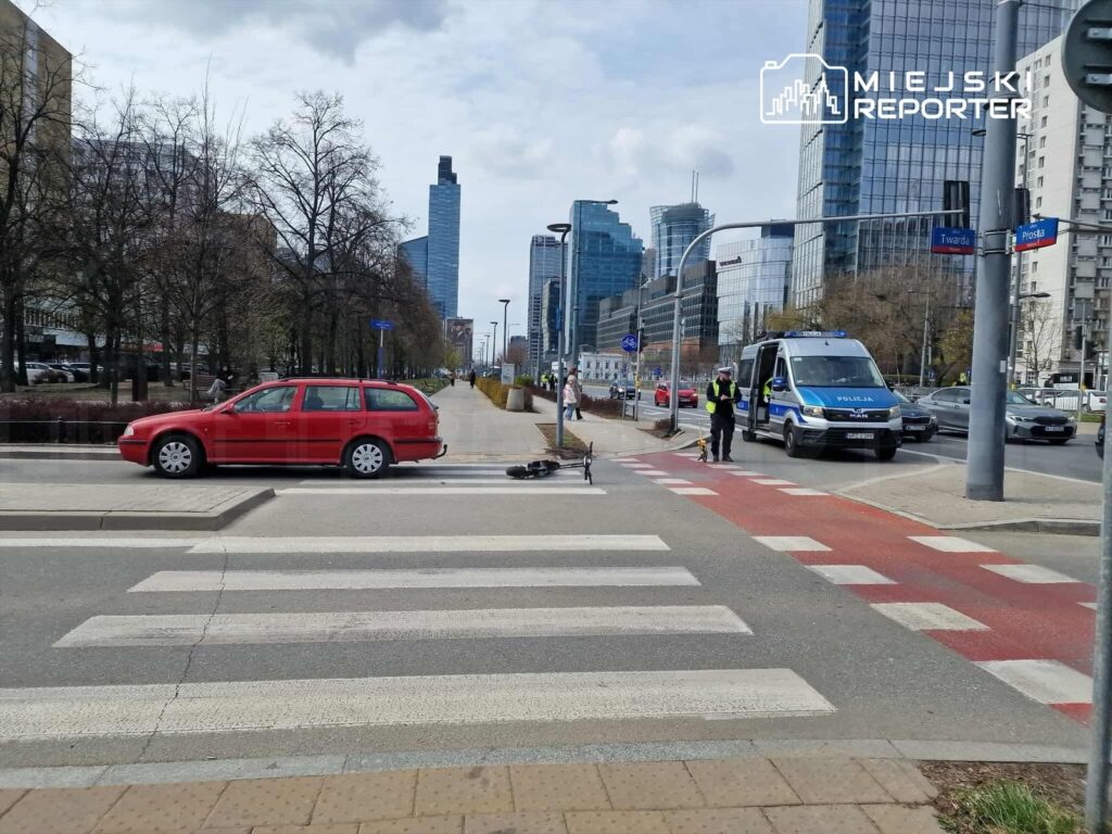 Czerwony samochód osobowy zatrzymany na przejściu dla pieszych, obok policjant z psem służbowym w miejskim otoczeniu.